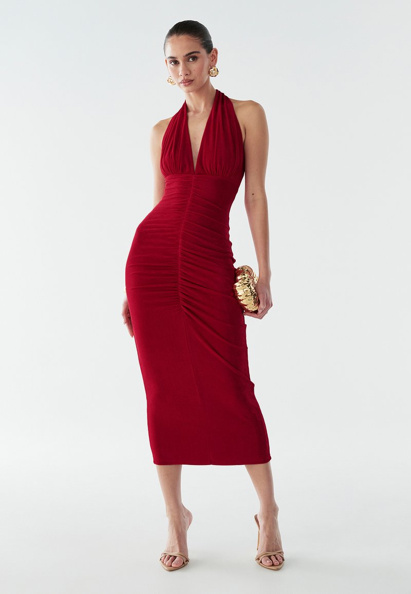 Raudona halterio suknele su raukšlėmis, prigludusi siluetas ir iki kelių ilgio. Modelis laiko auksinę rankinę ir užsima neutralias aukštakulnes batus.