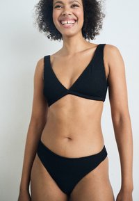 Juodas megztas bikinis, su V formos viršumi platesnėmis petnešėlėmis ir aukštu juosmeniu. Tekstūruota medžiaga be siūlių.