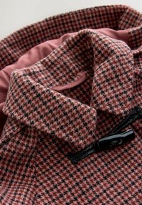 Villasegude segu, mis on kaetud roosa ja musta houndstooth mustriga, mustade nööbiga ja pehme roosa voodriga.