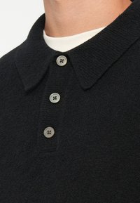 Schwarzes Poloshirt aus strukturiertem Stoff, mit einem Kragen und drei grauen Knöpfen auf der Vorderseite, darüber getragen über einem weißen Hemd.