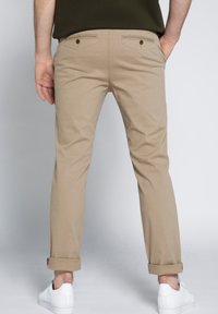 Chino beige chiaro con orli arrotolati, realizzati in tessuto di cotone. Due tasche posteriori con bottoni marroni e un design slim fit.