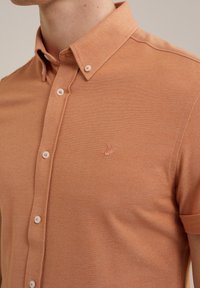 Krátký rukáv, světle oranžová košile na knoflíky s texturovanou tkaninou, která má límec a malou vyšitou nášivku na hrudi.