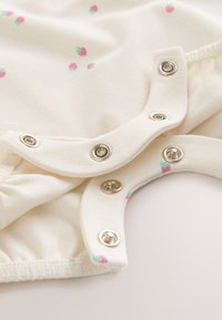 Närbild på krämfärgat babytyg med små rosa jordgubbsmönster och silverfärgade tryckknappar längs kanten.