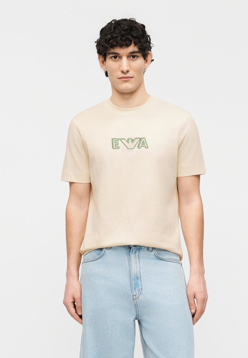 Νεαρός άνδρας με σγουρά μαλλιά, που φοράει ανοιχτό μπεζ μπλουζάκι με πράσινο λογότυπο "EVA" και ανοιχτό μπλε τζιν, στέκεται μπροστά σε λευκό φόντο.