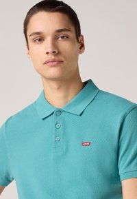 Tyrkysová polo košeľa Levi's s golierom, trojitou gombíkovou légiou a malým červeným logom na hrudi. Textúrová látka.