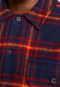 Camicia flanella blu navy con motivo a quadri rossi e arancioni, con chiusura abbottonata, colletto classico e taschino sul petto.