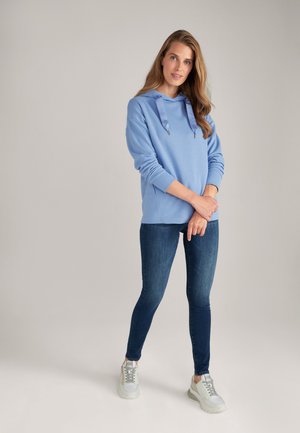 Lichtblauwe hoodie met een koord, gecombineerd met donkerblauwe skinny jeans en witte sneakers met grijze accenten. Glad textuur en casual ontwerp.