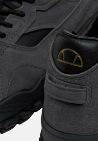 Sneaker in suede grigio con accenti neri, caratterizzati da una superficie testurizzata e un tallone nero con un logo dorato su un design arcuato.