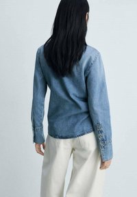 Femme aux longs cheveux noirs portant une veste en jean bleue et un pantalon blanc, debout de dos sur un fond uni.