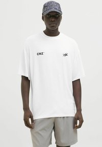 Balta per didelę T-marškiniai su juodu "CNZ" logotipu ir grafiniu akcentu, poruojama su šviesiai pilkomis šortais ir raštuota tamsia kepure.