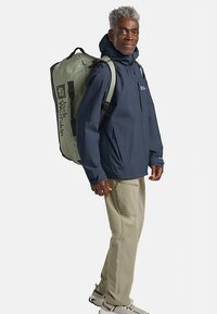 Wasserabweisende marineblaue Jacke mit verstellbarer Kapuze, beigen Hosen und grauem Rucksack mit Logo und Reißverschluss, getragen von einer stehenden Person.