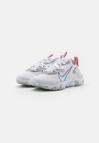 Zapatillas de malla blancas con acentos azules y detalles rojos, que cuentan con una parte superior texturizada, punta redonda y una suela flexible con un patrón.