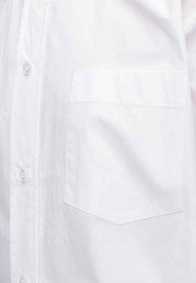 Chemise en coton blanc à l’avant avec une poche plaquée sur la poitrine gauche et des boutons blancs le long de la patte de boutonnage.