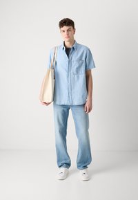 Camisa de mezclilla azul clara con mangas cortas, bolsillo en el pecho; combinada con jeans azul claros y zapatillas blancas; llevando una bolsa de mano crema.