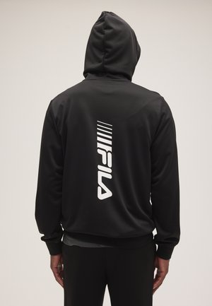 Persona que lleva una sudadera con capucha negra con el logo FILA y diseño a rayas blancas en el centro de la espalda, de espaldas a la cámara.