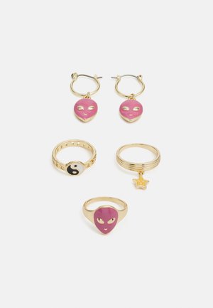 Conjunto de joias em tom dourado com argolas em forma de rosto alienígena rosa, um anel yin-yang e um anel com um encanto de estrela; acabamentos suaves e polidos.