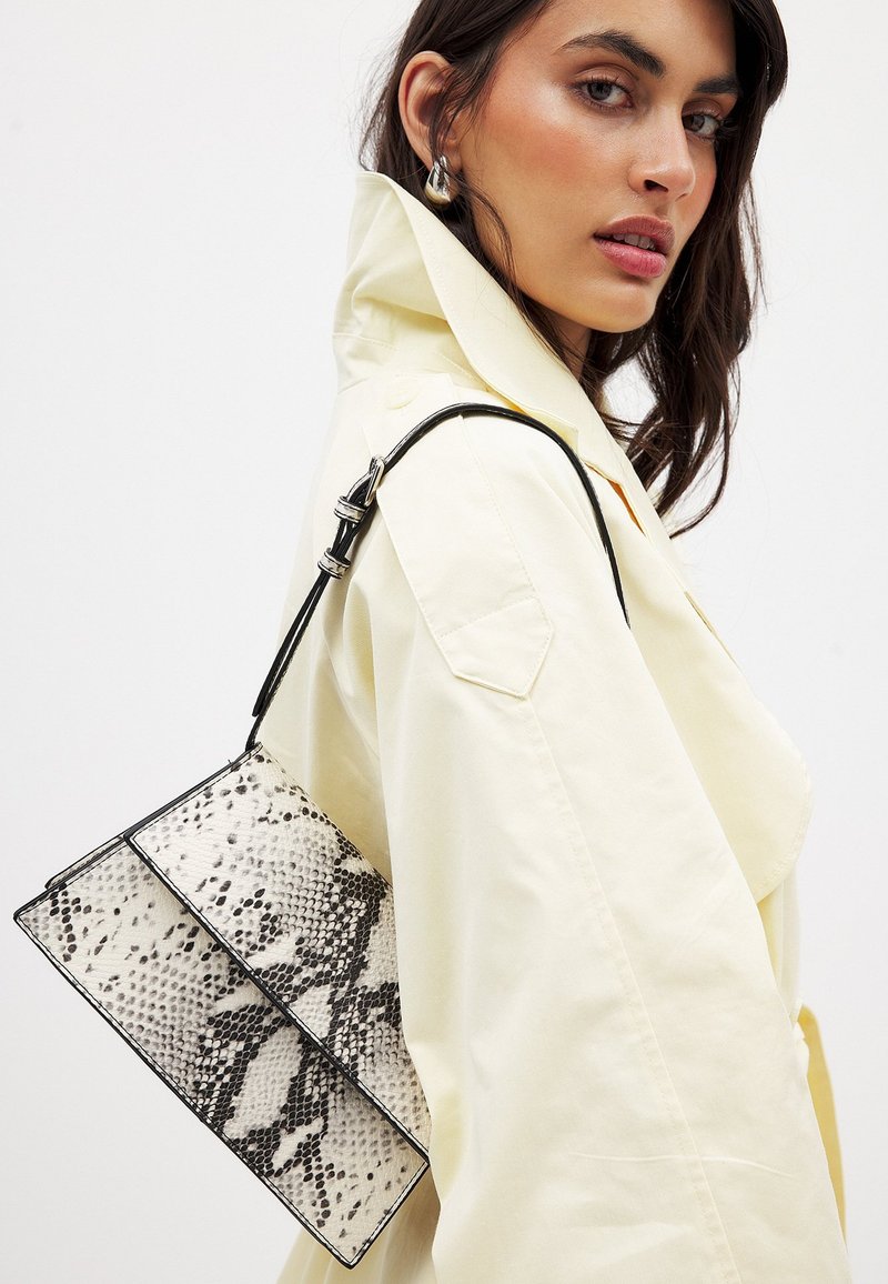 Črna in bela torbica s potiskom usnja kač, ki ima strukturirano obliko, z črnimi naramnicami in srebrnimi kovinskimi detajli.