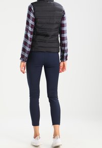 Czarna kamizelka puchowa nałożona na czerwoną i niebieską koszulę w kratę, w połączeniu z granatowymi dopasowanymi spodniami i białymi sneakersami; gładki materiał i elegancki krój.