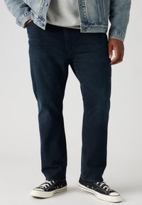 Donkerblauwe spijkerbroek met een rechte snit, met subtiele stikdetails en een versleten textuur. Gecombineerd met zwart-witte sneakers.