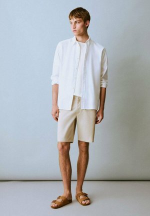 Chemise blanche à boutons, shorts beiges, sandales marron. Tissu léger, coupe décontractée et couleurs neutres. Pose debout sur un fond pâle.