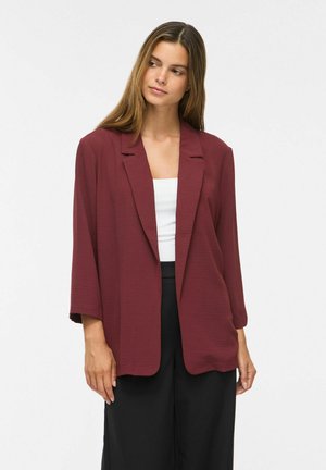 Femme aux longs cheveux bruns portant un blazer bordeaux ample par-dessus un haut blanc et un pantalon noir, debout devant un fond clair uni.