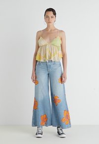 Top cropped em verde-amarelo com padrões, combinado com jeans de perna ampla azuis com grandes estampas florais em laranja. Ténis pretos completam o look.