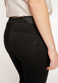 Vaqueros negros de mezclilla con un diseño ajustado, que cuentan con bolsillos traseros y una cintura texturizada con un logo. Superficie lisa y detalle clásico de costuras.