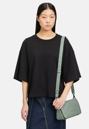 Kobieta o prostych ciemnych włosach, ubrana w luźną czarną koszulkę i jeansową spódnicę, trzymająca jasnosiwozieloną skórzaną torebkę na ramię.