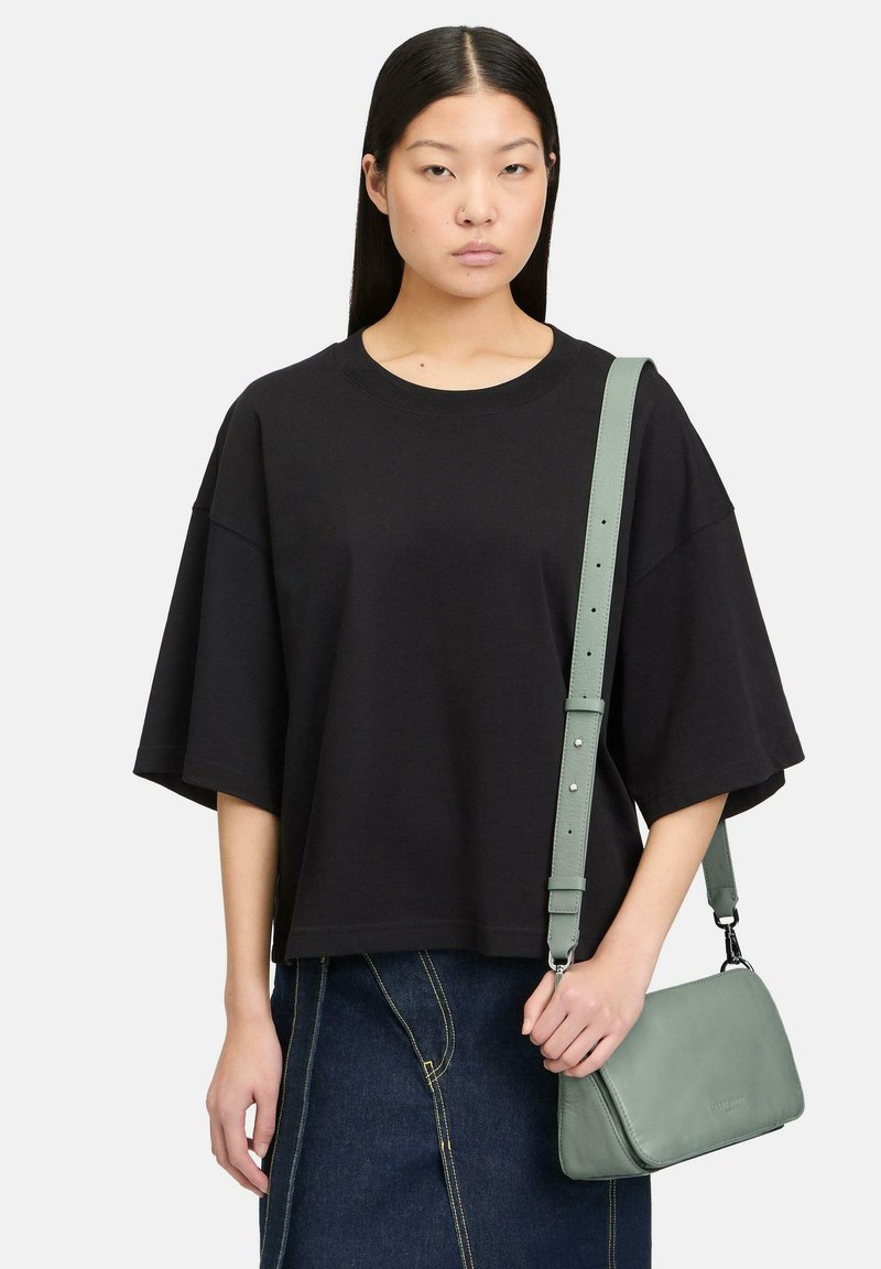 Kobieta o prostych ciemnych włosach, ubrana w luźną czarną koszulkę i jeansową spódnicę, trzymająca jasnosiwozieloną skórzaną torebkę na ramię.