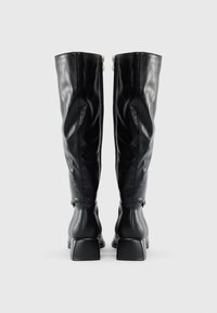 Un par de botas de cuero negras hasta la rodilla con tacones cuadrados y cremalleras en la parte posterior sobre un fondo blanco liso.