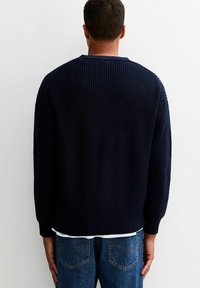 Marinblå stickad tröja med ribbat mönster och rundad halsringning, med nedhasade axlar och en lätt cropped nederkant, bärs över en vit skjorta.