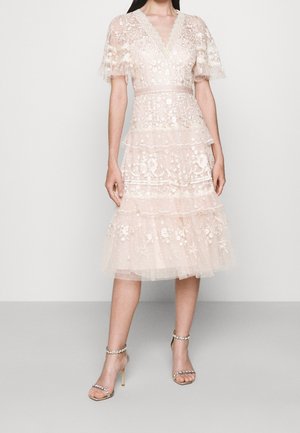 Femme portant une robe en dentelle transparente jusqu'aux genoux avec broderie florale et jupe à volants, associée à des talons ouverts avec brides à la cheville.