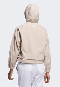 Ljusbeige huvjacka med elastiska ärmslut och nederkant, med en logotyp på baksidan. Tillverkad av ett mjukt, lätt tyg.