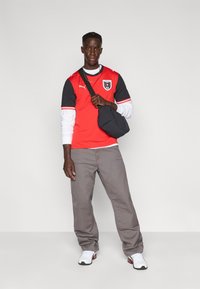 Vörös és fekete sportmez logóval, szürke nadrág, fekete táska, fehér hosszú ujjúval és fehér tornacipővel viselve. Egyszínű háttér.