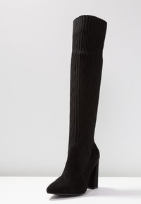 Botas negras hasta la rodilla con un upper de tela acanalada, punta afilada y tacón cuadrado. El material tiene una textura suave con un acabado liso.