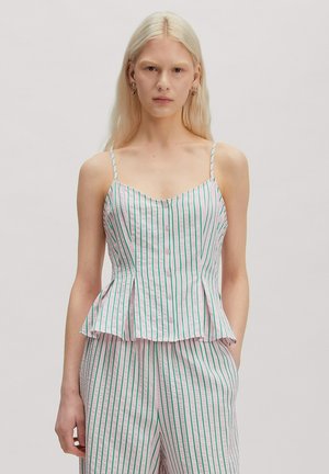 Blonde Frau trägt ein weißes ärmelloses, geknöpftes Oberteil und passende Hose mit grünen vertikalen Streifen und steht vor einem neutralen Hintergrund.