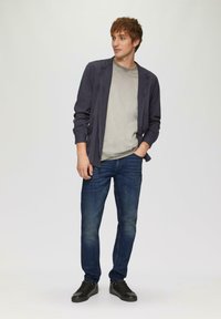 Camisa gris oscuro de botón sobre una camiseta gris claro, jeans ajustados de color azul y zapatillas negras. Atuendo casual sobre un fondo neutro.