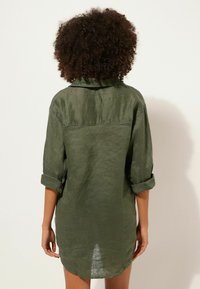 Camicia di lino verde oliva con colletto, maniche arrotolate e orlo curvo. Presenta una spalla posteriore e una vestibilità rilassata.