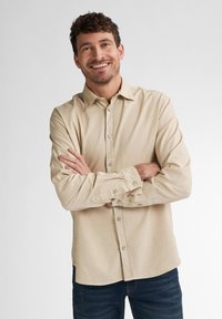 Beige blouse met lange mouwen, knopenrij aan de voorkant en manchetten met één knoop, gemaakt van een gladde stof, gedragen met donkerblauwe jeans.