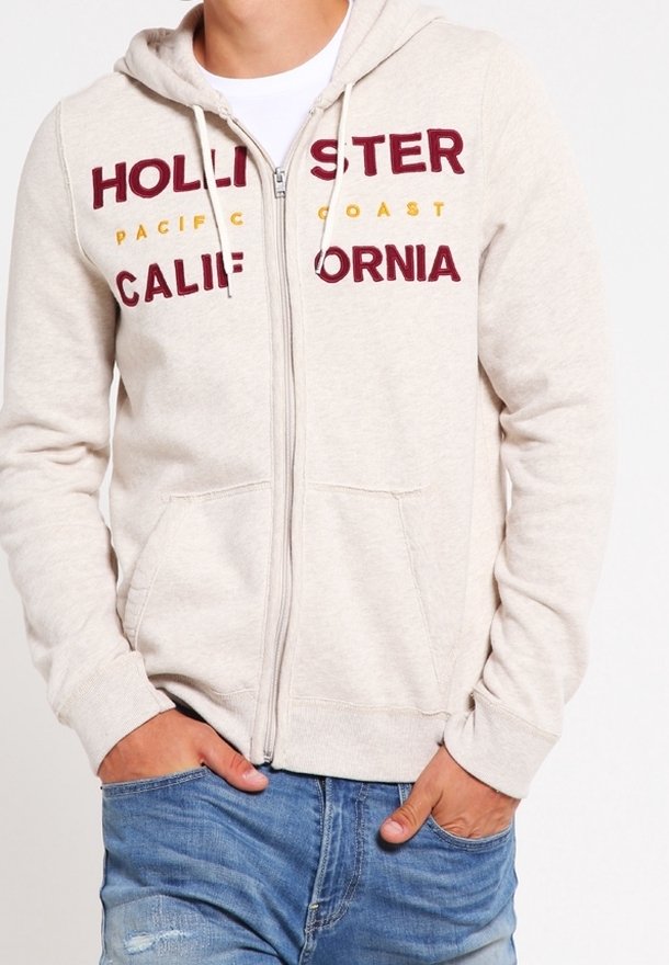 Sweat-shirt à fermeture éclair à capuche en mélange de coton beige clair. Présente un texte imprimé rouge et jaune "HOLLISTER PACIFIC COAST CALIFORNIA" et des poches frontales.