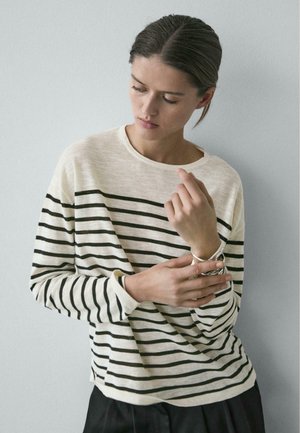 Femme portant un chemisier à manches longues rayé blanc et noir ajustant un bracelet argenté à son poignet gauche sur un fond gris clair uni.