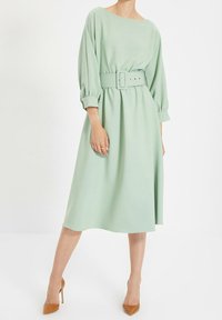 Miętowo-zielona sukienka do kolan z okrągłym dekoltem, długimi bufkami i dopasowanym paskiem. Miękka, gładka faktura; prosty design.