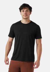 Mand med kort krøllet hår iført en ensfarvet sort kortærmet t-shirt og brune bukser, stående mod en hvid baggrund.