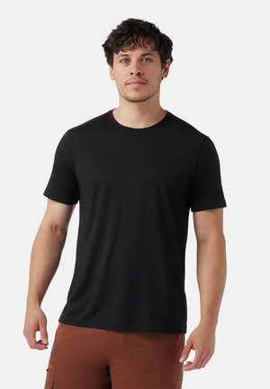 Mann mit kurzem lockigem Haar, der ein einfaches schwarzes Kurzarm-T-Shirt und braune Hosen trägt, steht vor einem weißen Hintergrund.