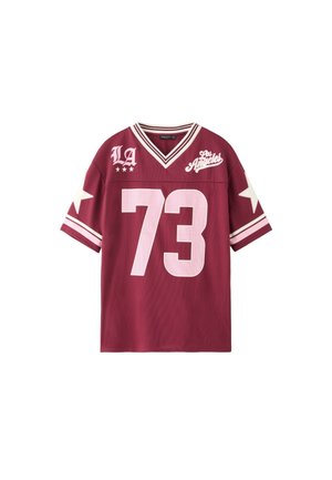 Jeune femme aux longs cheveux portant un maillot de sport bordeaux avec le numéro 73, un pantalon cargo ample et des bottes marron sur fond vert.