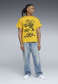 Gul bomulds T-shirt med en sort grafik af en bi, der holder en fodbold, parret med lyseblå løstsiddende denim jeans og hvide sneakers.