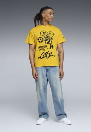 Gul bomulds T-shirt med en sort grafik af en bi, der holder en fodbold, parret med lyseblå løstsiddende denim jeans og hvide sneakers.