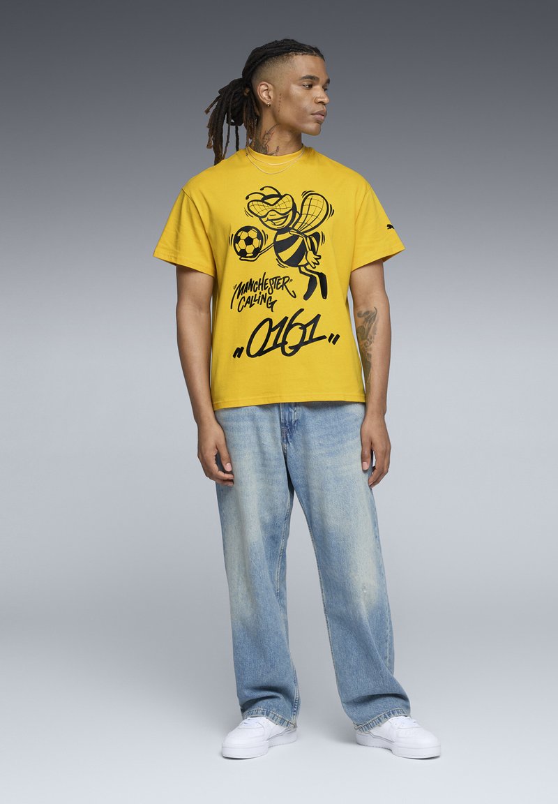 Gul bomulds T-shirt med en sort grafik af en bi, der holder en fodbold, parret med lyseblå løstsiddende denim jeans og hvide sneakers.