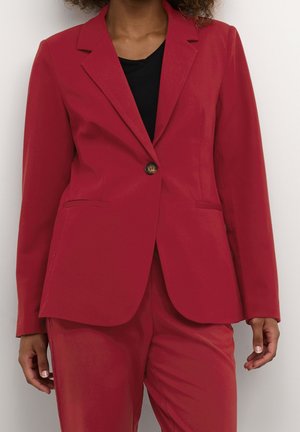 Woman wearing a tailored red blazer with one button, paired with matching red trousers and a black top underneath.