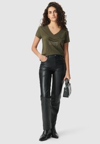 Camiseta verde oliva con cuello en V con "1987" impreso en negro, combinada con pantalones de cuero negro de cintura alta y un pequeño bolso negro.